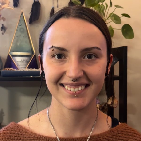 Young woman with a smile, featuring facial piercings and a brown sweater.