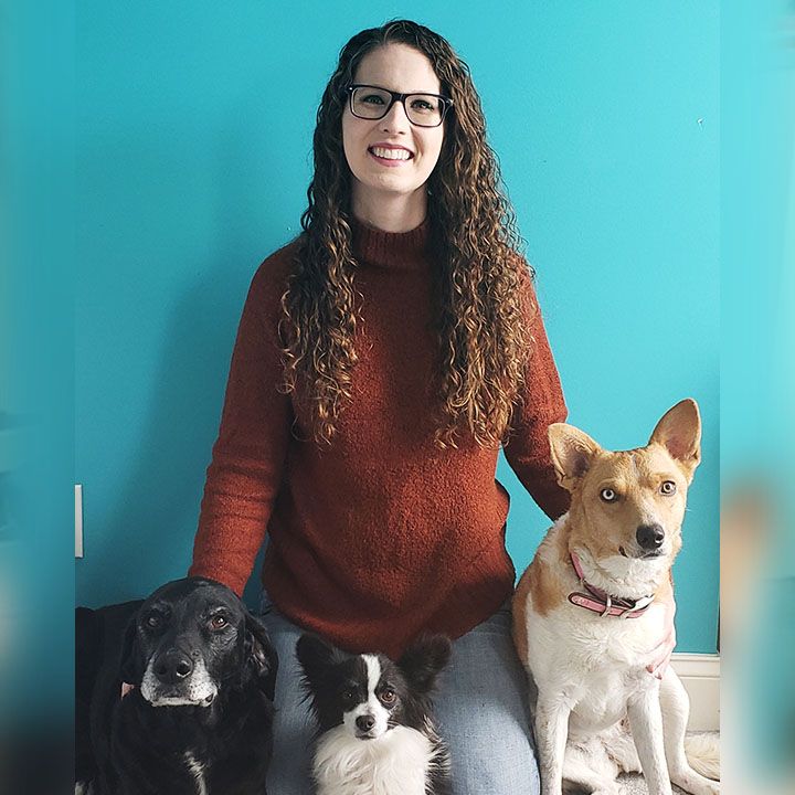Woman with glasses and curly hair, smiling, seated with three dogs of varied breeds.