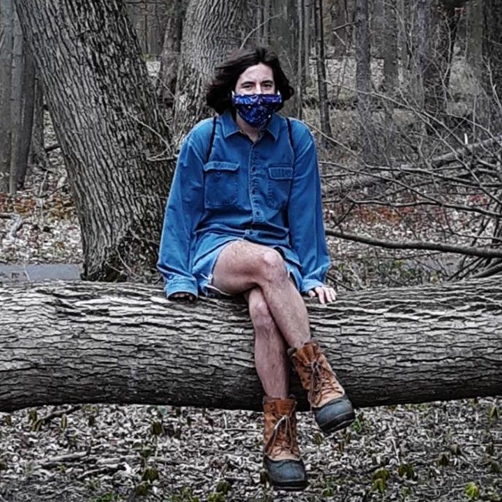 Woman in blue sitting on a fallen tree wearing a mask and boots.
